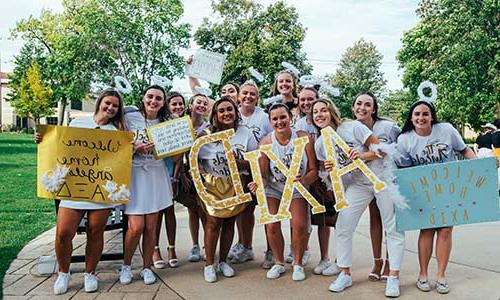A group of smiling young women dressed in white with angel-themed accessories, holding signs and letters that spell out "axid," possibly welcoming new members to their sorority.